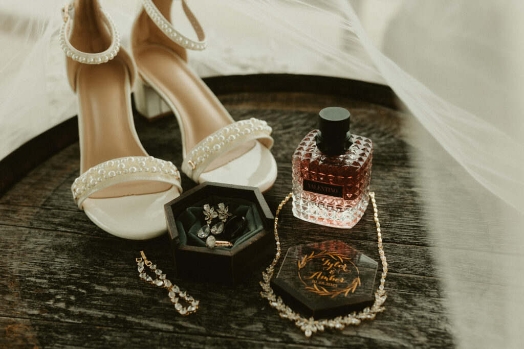 Wedding detail photo containing the bride's shoes, ring box, earrings, bracelet, necklace, and perfume.